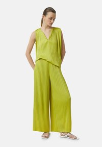 Limoengroene plooijumpsuit met een V-hals, wijde broekspijpen en lichtgewicht stof, gestyled met eenvoudige witte sandalen.