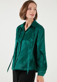 Emerald green satin blouse with a pointed collar, long sleeves, and side ties. Features smooth texture and a button-down front design.