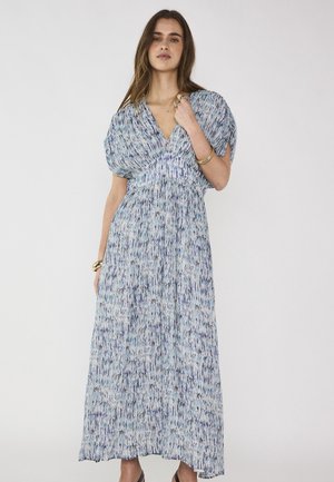 Jeune femme portant une robe longue bleu et blanc à motifs avec des manches bouffantes, debout devant un fond clair uni.