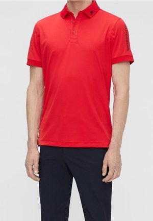 Homme portant un polo rouge vif à manches courtes avec un texte noir sur la manche gauche et un pantalon foncé sur un fond clair uni.