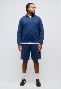 Άνδρας με πλεγμένα μαλλιά, φοράει μπλε σκούρο φούτερ με φερμουάρ στο στήθος, ταιριαστά σορτς, λευκές κάλτσες και μαύρα αθλητικά παπούτσια, στέκεται και κοιτάζει δεξιά.