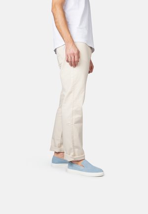 Persona con pantalones beige arremangados, camisa blanca y zapatos tipo slip-on azul claro, de pie contra un fondo claro y liso.