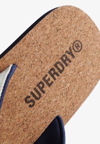 Piano in sughero con logo "SUPERDRY®" impresso in nero, bordato da una tracolla in tessuto blu scuro. Design solido e texture.