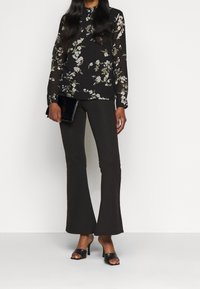 Femme portant un chemisier noir à fleurs avec des manches transparentes, un pantalon noir évasé, des sandales à talons noires, et tenant une pochette noire.