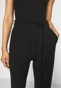 Mono negro con corpete ajustado, piernas anchas y un cinturón atado a la cintura. Textura suave y diseño minimalista, con bolsillos laterales.