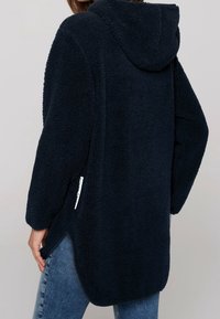 Marinblå huvtröja i fleece med texturerad tyg, sidofickor och en etikettdetalj vid fållen, med avslappnad passform och sänkta axlar.