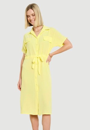 Uśmiechnięta kobieta z blond bobem w żółtej sukience na guziki do kolan z krótkimi rękawami i paskiem w pasie, stojąca na jednolitym tle.