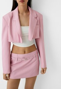 Blazer corto rosa con rever a punte, abbinato a una mini gonna coordinata con cintura e hardware argentato. Top bianco aderente sotto.