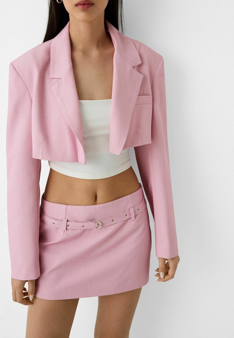 Blazer corto rosa con rever a punte, abbinato a una mini gonna coordinata con cintura e hardware argentato. Top bianco aderente sotto.