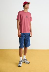 T-shirt en coton rose courte avec un logo Timberland noir, associé à un short en denim bleu et des chaussures de sport grises, se tenant sur un sol jaune.