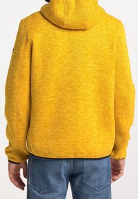 Gul huvtröja med en texturerad yta och huva. Funktioner inkluderar sidofickor och en mörk nederkant. Bärs med blå jeans.