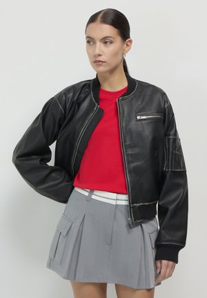 Veste bomber en cuir noir avec des poignets côtelés, une poche latérale et une poche zippée. Associée à une chemise rouge et une jupe plissée grise.