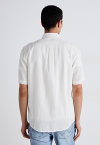 Camicia bianca in lino a maniche corte con colletto standard, piega sul retro e orlo curvo, abbinata a jeans azzurri chiari.