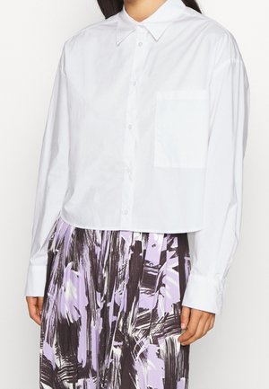 Kobieta ubrana w luźną białą koszulę zapinaną na guziki z kieszonką na piersi oraz plisowaną spódnicę z czarnymi i lawendowymi abstrakcyjnymi wzorami.