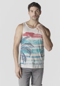 Uomo che indossa una canottiera bianca con strisce di palme rosse, blu e verde-azzurro, e pantaloncini grigio chiaro, in piedi davanti a uno sfondo semplice.