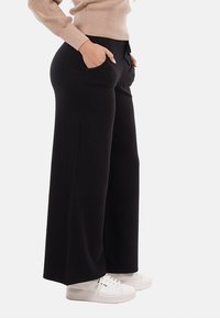 Persona che indossa pantaloni neri a gamba larga con le mani in tasca, maglione beige e sneakers bianche su uno sfondo bianco.