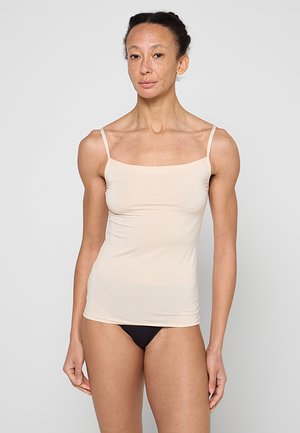 Frau mit zurückgebundenen Haaren, die ein beiges Camisole und schwarze Unterwäsche trägt, steht vor einem schlichten hellgrauen Hintergrund.