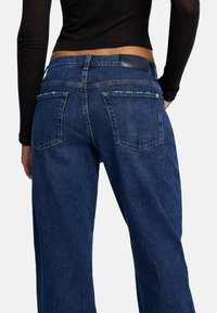 Jeans di denim blu scuro con vestibilità dritta. Presentano due tasche posteriori e una patch in pelle sul girovita. La texture appare leggermente robusta.