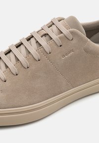 Baskets en suède beige avec des lacets ronds plats, une semelle en caoutchouc lisse et un logo en relief sur le côté. Bout arrondi et détails de conception minimalistes.