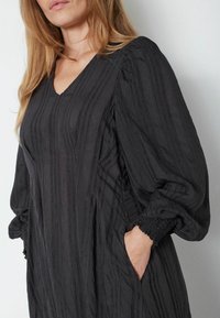 Čierne šaty s výstrihom do V, dlhými puff rukávmi s elastickými manžetami a textúrovaným vzorom s vertikálnymi pruhmi. Viditeľné sú dve bočné vrecká.