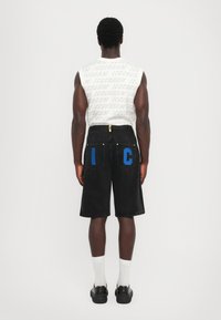 Pantalones cortos de mezclilla negra con letras gráficas azules "I" y "C" en los bolsillos traseros. Combinados con una camiseta sin mangas blanca y zapatos negros.