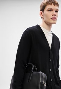 Cardigã preto com decote em V, com botões na parte da frente. Combinado com uma t-shirt branca e segurando uma bolsa de couro preta.