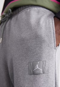 Grå mjukisbyxor med en synlig Jumpman-logotyp på fickan, bärs med ett färgglatt klädlager ovanpå.