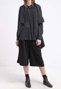 Czarny zestaw składający się z długiej, otwartej kurtki, plisowanej bluzki i szerokich spodni typu culottes. Wykonany z lekkiej tkaniny o minimalnej fakturze.