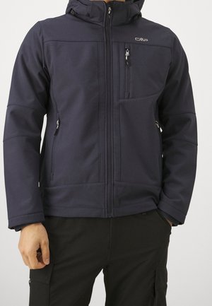 Mężczyzna w ciemnoniebieskiej kurtce na zamek błyskawiczny z kapturem oraz kieszeniami na zamek na piersi i po bokach, w połączeniu z czarnymi spodniami bojówkami.