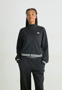 Schwarze Sportjacke mit hohem Kragen und durchgehendem Reißverschluss, ausgestattet mit weißen gestreiften Akzenten an den Bündchen und am Saum, kombiniert mit passenden Hosen.