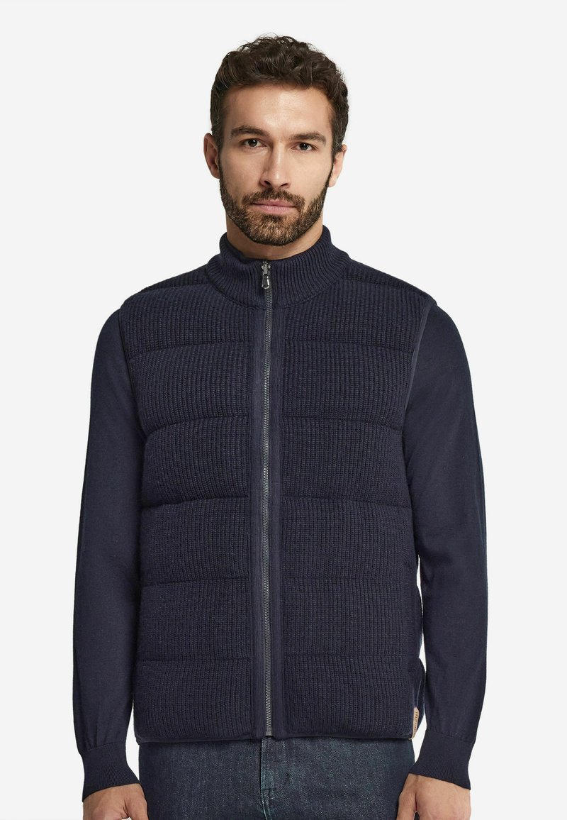 Mörkblå stickad väst med vertikal ribbad textur, hög krage och framficksöppning; bärs över en långärmad mörk skjorta.