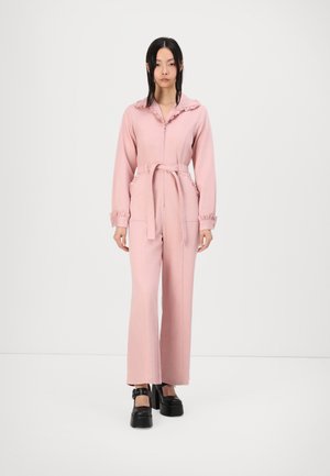 Salopette rose avec col et poignets volantés, taille ceinturée, manches longues et poches latérales ; le tissu semble lisse et léger. Chaussures à plateforme noires.