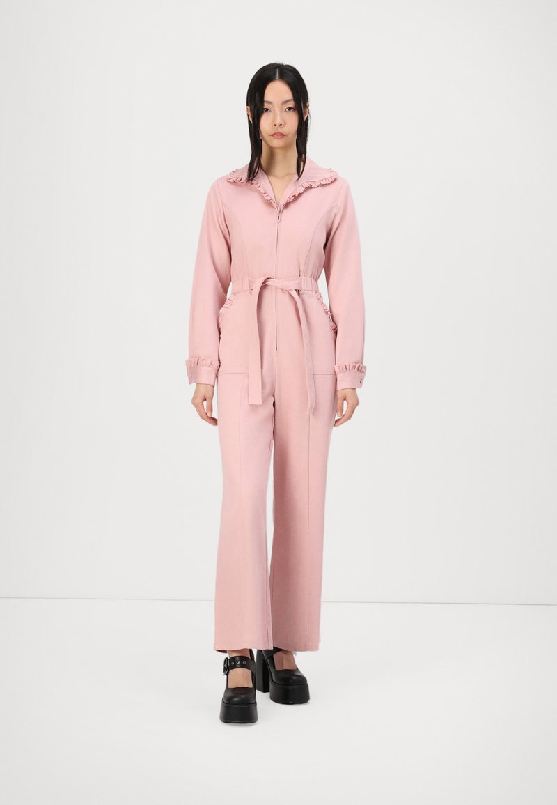 Macacão rosa com colarinho e punhos de babados, cintura cevada, mangas longas e bolsos laterais; o tecido parece liso e leve. Sapatos de plataforma pretos.