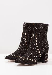 Mustat mokkasiinimaiset nilkkakengät, joissa on kultaiset niittiyksityiskohdat, kärjen muotoilu on terävä ja kantapää on paksu. Takana vetoketjukiinnitys.