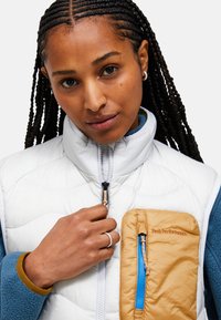 Vit quiltad väst med hög krage, med en beige dragkedjeficka och blå accenter. Ytan är slät med synliga sömdetaljer.