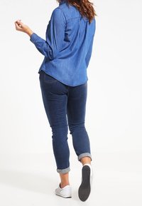 Jeansskjorta i medelblått med långa ärmar och rundad nederkant, i kombination med mörkblå jeans, uppvikta ärmslut och vita sneakers.