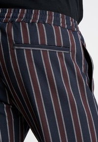 Pantalons rayés bleu marine et bordeaux avec un tissu texturé, dotés d'une taille élastique et d'une poche arrière avec une ouverture horizontale.
