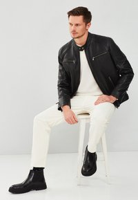 Zwarte leren jas met ritszakken, witte trui en witte broek. Zwarte Chelsea-boots met tekstuurlijke zool, zittend op een kruk.