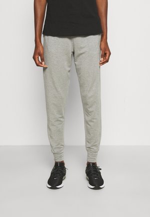 Person iført grå joggerbukser og sorte sneakers, stående mod en ensfarvet lys baggrund med hænder afslappet ved siden.