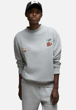 Personne portant un sweat-shirt gris avec une petite photo imprimée et une poche boutonnée, un pantalon gris assorti et une casquette noire et blanche.