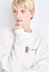 Balts sporta džemperis ar ribotām piedurknēm, kuram ir krāsains izšūts logo un melna uzraksts "TOMMY JEANS". Mīksta tekstūra, brīvs piegriezums.