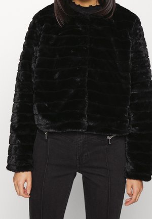 Vrouw draagt een zwart cropped faux fur-jasje met lange mouwen en zwarte hoge taillebroeken met ritssluiters.