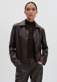 Braune Kunstlederjacke mit Kragen, zwei Knöpfen und Seitentaschen, kombiniert mit einem passenden braunen Oberteil und einer Hose.
