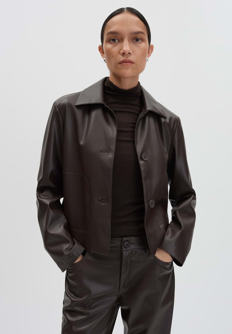 Braune Kunstlederjacke mit Kragen, zwei Knöpfen und Seitentaschen, kombiniert mit einem passenden braunen Oberteil und einer Hose.