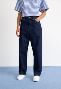 Jean en denim bleu foncé, à jambes larges, avec des coutures jaunes et des poches avant, associés à une chemise rayée bleu clair et blanche. Baskets blanches.