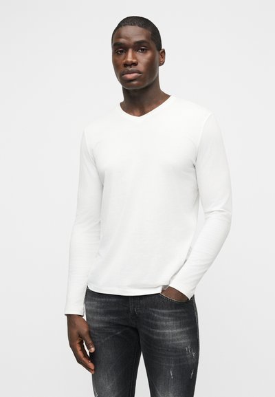 Άνδρας που φοράει απλό λευκό μακρυμάνικο πουκάμισο και σκουρόχρωμα τζιν με φθορές, στέκεται με το ένα χέρι στην τσέπη μπροστά σε φωτεινό φόντο.