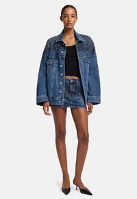 Giacca di jeans con design a gradient di borchie, maniche lunghe, vestibilità oversize; abbinata a una gonna di jeans coordinata e tacchi neri.