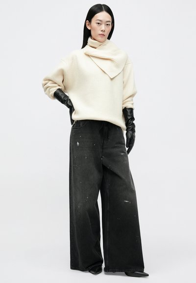 Puhasta krema pulover z rebrasto teksturo in asymetrično ovratno podrobje, v kombinaciji z širokima črnima kavbojkami z madeži barve in usnjenimi rokavicami.