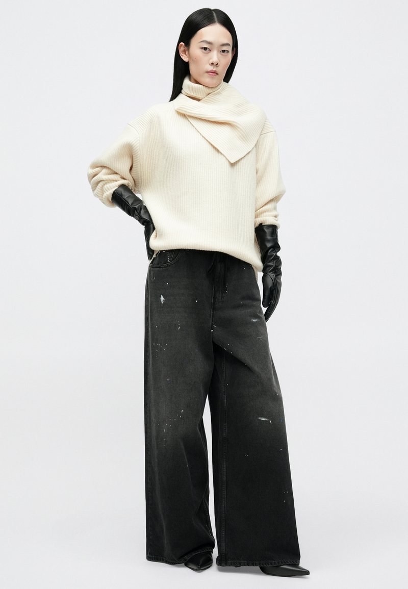Chunky crème trui met ribbeltextuur en asymmetrische nekdetail, gecombineerd met wijdlopende zwarte jeans met verfspetters en leren handschoenen.