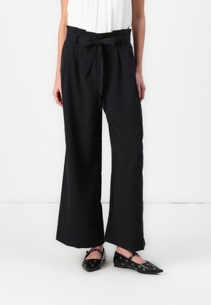 Mujer que lleva pantalones negros de pierna ancha y cintura alta con un cinturón atado, y zapatos planos negros con punta puntiaguda y cortes circulares.